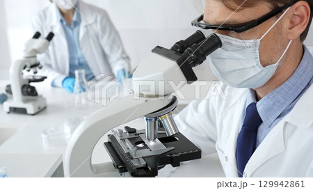Masked scientist analyzing sample under microscope, colleague researching nearby in sterile medical laboratory setting. Medicine, healthcare and science concept Masked scientist analyzing sample under microscope, colleague researching nearby in sterile medical laboratory setting. Medicine, healthcare and science concept 129942861