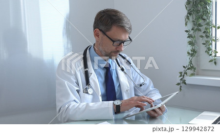 Medical doctor wearing lab coat and stethoscope using digital tablet consulting patient data in bright medical office with laptop next to him. Medicine and health care 129942872