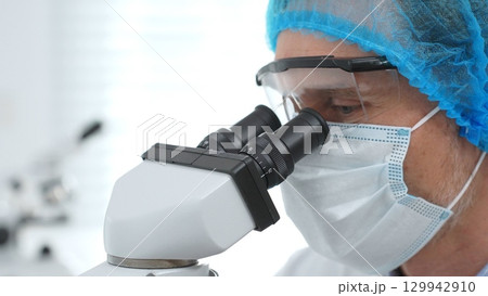 Masked scientist examining sample through microscope, wearing protective lab gear during medical research. Medicine, healthcare and science concept Masked scientist examining sample through microscope, wearing protective lab gear during medical research. Medicine, healthcare and science concept 129942910