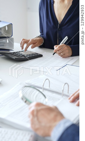 Business colleagues are reviewing financial reports and using a calculator during a productive meeting in a brightly lit office setting. Audit and taxes in business 129942933