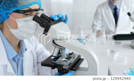 Female scientist wearing a mask and blue safety gear, using a microscope for research while a male colleague works in the background. Medicine, healthcare and science concept 129942940