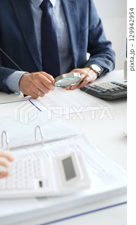 Male financial analysts examining documents, using magnifying glass and calculator while studying corporate data at workplace. Audit and taxes concept Male financial analysts examining documents, using magnifying glass and calculator while studying corporate data at workplace. Audit and taxes concept 129942954