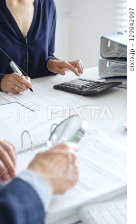 Female financial analysts examining documents, using magnifying glass and calculator while studying corporate data at workplace. Audit and taxes 129942997