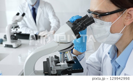 A female scientist in a blue mask and gloves is using a microscope and conducting scientific research in a sterile laboratory while a colleague is working in the background. Medicine, healthcare and 129942998