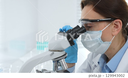 Female scientist wearing a mask and protective glasses using a microscope in a laboratory setting, conducting scientific research and analysis. Medicine, healthcare and science concept Female scientist wearing a mask and protective glasses using a microscope in a laboratory setting, conducting scientific research and analysis. Medicine, healthcare and science concept 129943002