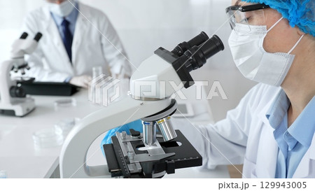 Female scientist wearing a mask and blue safety gear, using a microscope for research while a male colleague works in the background. Medicine, healthcare and science concept 129943005