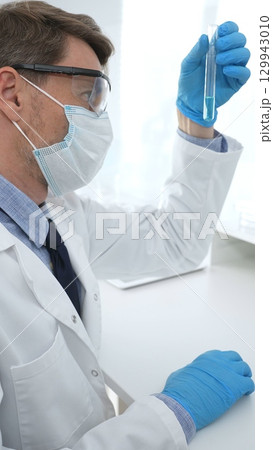 Scientist in a white lab coat, face mask, and gloves, analyzing a blue liquid in a test tube within a laboratory setting. Medicine, healthcare and science concept Scientist in a white lab coat, face mask, and gloves, analyzing a blue liquid in a test tube within a laboratory setting. Medicine, healthcare and science concept 129943010
