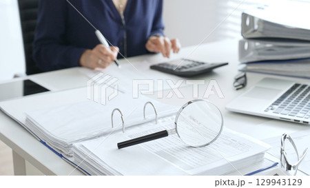 Magnifying glass and calculator are on the table over financial documents opposite female accountant analyzing data, reviewing reports, binders and laptop on a white desk. Audit and taxes concept Magnifying glass and calculator are on the table over financial documents opposite female accountant analyzing data, reviewing reports, binders and laptop on a white desk. Audit and taxes concept 129943129