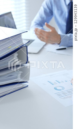 Corporate professionals reviewing financial documents, working collaboratively at white desk with blue folder stack, highlighting teamwork and business productivity. Audit and taxes in business Corporate professionals reviewing financial documents, working collaboratively at white desk with blue folder stack, highlighting teamwork and business productivity. Audit and taxes in business 129943139
