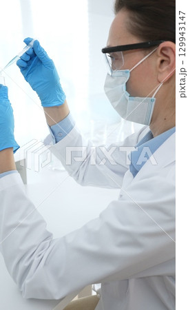 Female scientist wearing a face mask and blue gloves, carefully dropping liquid from a pipette into a test tube in a modern lab. Medicine, healthcare and science concept 129943147