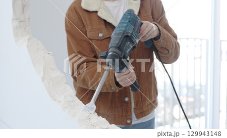 Unrecognizable construction worker demolishing concrete wall with rotary hammer, wearing protective safety gear. Renovation concept, closeup vertical view 129943148
