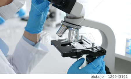 Close up of scientific professional blue gloved hands examining microscope lens, performing detailed medical laboratory research. Medicine, healthcare and science concept Close up of scientific professional blue gloved hands examining microscope lens, performing detailed medical laboratory research. Medicine, healthcare and science concept 129943161