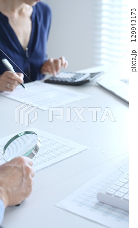 Two professional accountants analyzing financial reports with magnifying glass and calculator, performing accounting and auditing tasks in the office, close up of hands. Audit and taxes in business Two professional accountants analyzing financial reports with magnifying glass and calculator, performing accounting and auditing tasks in the office, close up of hands. Audit and taxes in business 129943173