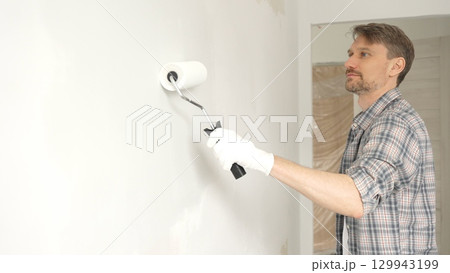 Man professional painter wearing checkered shirt, is rolling white paint on apartment wall with paint roller, transforming interior space with steady strokes during comprehensive home renovation 129943199