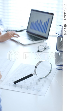 Close up of magnify glass over financial documents near blue folders full of financial papers, accountants are analyzing reports and documents at the background. Audit and taxes in business Close up of magnify glass over financial documents near blue folders full of financial papers, accountants are analyzing reports and documents at the background. Audit and taxes in business 129943227
