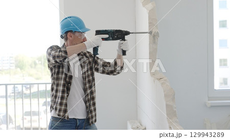 Man construction worker demolishing a wall with a hammer drill in protective gear, creating construction debris on the surrounding floor surface. Renovation concept 129943289