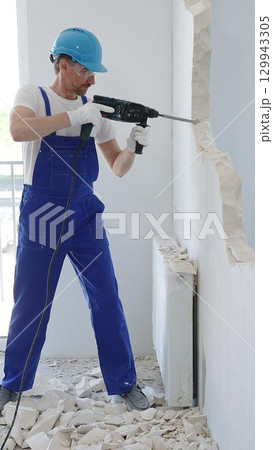 Man construction worker demolishing concrete wall with rotary hammer, wearing protective safety gear, generating dust and debris during home renovation work Man construction worker demolishing concrete wall with rotary hammer, wearing protective safety gear, generating dust and debris during home renovation work 129943305