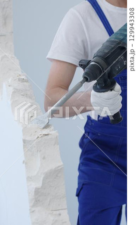 Construction worker demolishing white wall with rotary hammer drill, wearing blue overalls and protective white gloves, generating dust during renovation project, closeup view 129943308