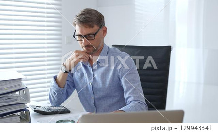 Serious male accountant is thinking while analyzing tax documents, using calculator near stacked folders, sitting in black office chair wearing crisp blue shirt. Audit and taxes in business Serious male accountant is thinking while analyzing tax documents, using calculator near stacked folders, sitting in black office chair wearing crisp blue shirt. Audit and taxes in business 129943341