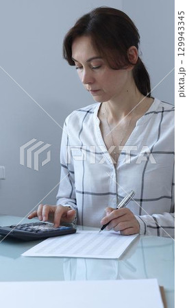 A professional woman calculates finances, checks documents, and accurately and carefully tracks business expenses at a glass table. Audit and taxes in business. A professional woman calculates finances, checks documents, and accurately and carefully tracks business expenses at a glass table. Audit and taxes in business. 129943345