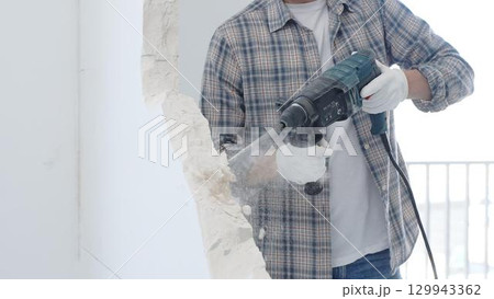 Male construction worker demolishing white wall with rotary hammer drill is wearing grey checkered shirt and protective white gloves, generating dust during renovation project, closeup view 129943362