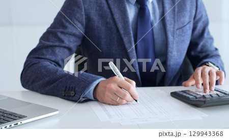 Businessman wearing suit and tie is using calculator and taking notes while working at desk in office, with laptop and stack of folders nearby, close up. Audit and taxes theme in business Businessman wearing suit and tie is using calculator and taking notes while working at desk in office, with laptop and stack of folders nearby, close up. Audit and taxes theme in business 129943368