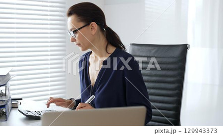 Professional female wearing black glasses, is working at desk, calculating finances, writing notes, managing business documents in contemporary workspace. Audit and taxes in business Professional female wearing black glasses, is working at desk, calculating finances, writing notes, managing business documents in contemporary workspace. Audit and taxes in business 129943373