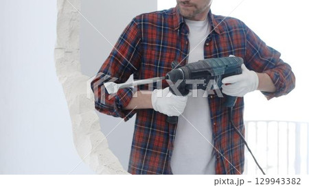 Male construction worker wearing red checkered shirt and protective white gloves is demolishing white wall with rotary hammer drill and generating dust during renovation project, closeup view 129943382