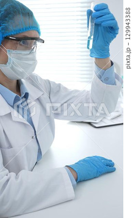 Medical science researcher wearing blue protective gear of cap, gloves and googles, is examining liquid in test tube in laboratory workspace. Medicine and health care concept Medical science researcher wearing blue protective gear of cap, gloves and googles, is examining liquid in test tube in laboratory workspace. Medicine and health care concept 129943388