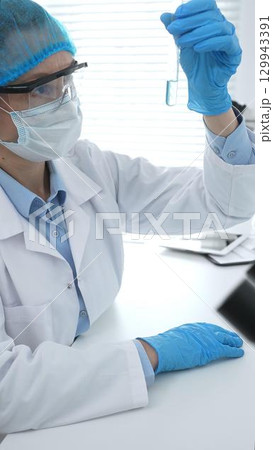 Medical science researcher wearing blue protective gear of cap, gloves and googles, is examining liquid in test tube in laboratory workspace. Medicine and health care concept Medical science researcher wearing blue protective gear of cap, gloves and googles, is examining liquid in test tube in laboratory workspace. Medicine and health care concept 129943391