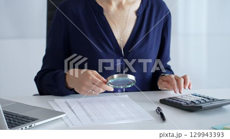Professional female accountant analyzing financial documents, using magnifying glass and calculator at workplace desk, focused on detailed accounting review, close up. Audit and taxes in business 129943393