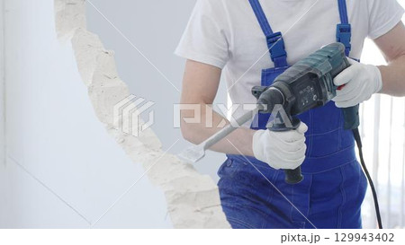 Construction worker demolishing white wall with rotary hammer drill, wearing blue overalls and protective white gloves, generating dust during renovation project, closeup view 129943402