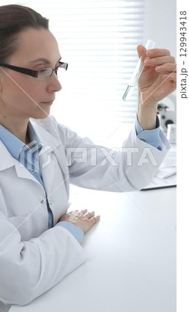 Female researcher examining clear liquid in test tube, sitting near microscopes and lab equipment within sterile scientific workspace. Medicine, healthcare and science concept 129943418