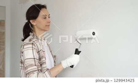 Middle aged woman wearing gloves is skillfully applying paint to a white wall with a paint roller in an apartment undergoing renovation, transforming the space with her creative touch Middle aged woman wearing gloves is skillfully applying paint to a white wall with a paint roller in an apartment undergoing renovation, transforming the space with her creative touch 129943425