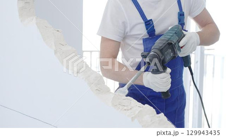 Male construction worker demolishing white wall with rotary hammer drill, wearing blue overalls and protective white gloves, closeup view 129943435