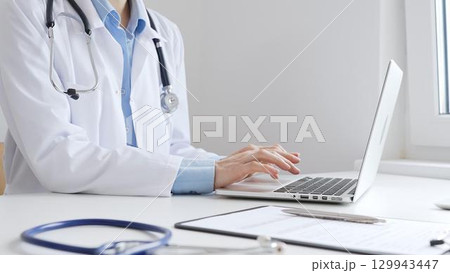 Doctor woman is typing on a laptop with a stethoscope, medical record, and pen on the desk in a bright medical office, showcasing healthcare technology and medical documentation. Medicine concept Doctor woman is typing on a laptop with a stethoscope, medical record, and pen on the desk in a bright medical office, showcasing healthcare technology and medical documentation. Medicine concept 129943447