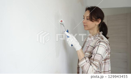 Woman wearing protective gloves painting white wall with small paint roller, updating interior and giving room clean, modern look during home renovation project 129943455