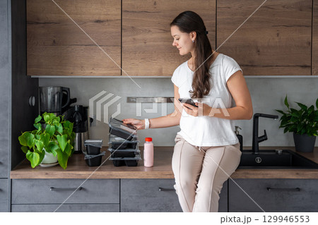 Young woman with healthy diet plan for weight loss, delivery of daily ready meal menu. Woman using meal tracker app on phone. Balanced portion with dish. Pre-cooking. Young woman with healthy diet plan for weight loss, delivery of daily ready meal menu. Woman using meal tracker app on phone. Balanced portion with dish. Pre-cooking. 129946553