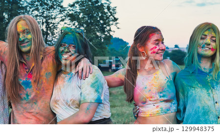 Four happy school friends hugging and smiling, covered in colorful holi powder paint, enjoying a summer outdoor fest 129948375