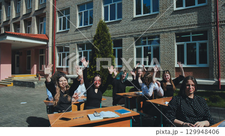 Happy students raising their hands at outdoor desks, having a great time during their school day Happy students raising their hands at outdoor desks, having a great time during their school day 129948406