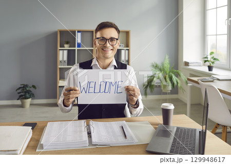 Man sitting at the desk in office and holding whiteboard with a message word welcome. Man sitting at the desk in office and holding whiteboard with a message word welcome. 129948617
