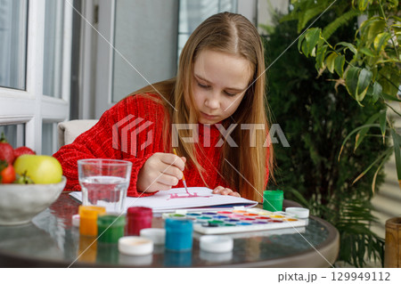 Portrait of young girl in red warm sweater drawing in a sketchbook while sitting on the balcony 129949112