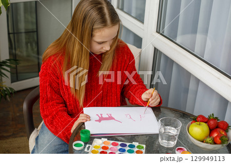 Attractive young girl in red warm sweater drawing in a sketchbook while sitting on the balcony 129949113