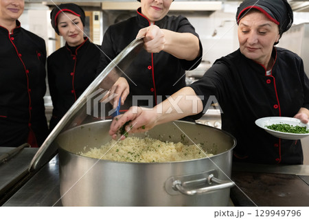 Group of chefs in black uniforms preparing rice in a large pot in a commercial kitchen. Female cooks stirring and adding herbs while working as a team. Concept of teamwork, gastronomy, and hospitality Group of chefs in black uniforms preparing rice in a large pot in a commercial kitchen. Female cooks stirring and adding herbs while working as a team. Concept of teamwork, gastronomy, and hospitality 129949796