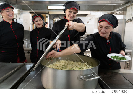 Group of chefs in black uniforms preparing rice in a large pot in a commercial kitchen. Female cooks stirring and adding herbs while working as a team. Concept of teamwork, gastronomy, and hospitality 129949802