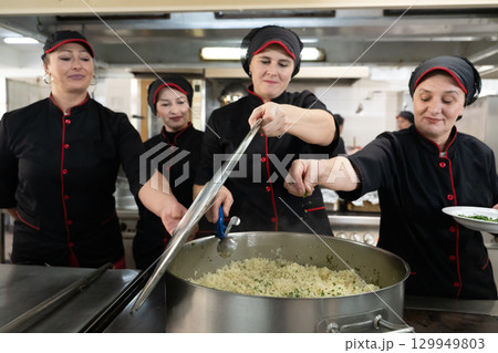 Group of chefs in black uniforms preparing rice in a large pot in a commercial kitchen. Female cooks stirring and adding herbs while working as a team. Concept of teamwork, gastronomy, and hospitality Group of chefs in black uniforms preparing rice in a large pot in a commercial kitchen. Female cooks stirring and adding herbs while working as a team. Concept of teamwork, gastronomy, and hospitality 129949803
