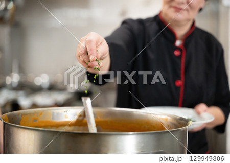 Close up of a chef in black uniform adding fresh herbs to a large pot of soup in a professional kitchen. Concept of gastronomy, seasoning, culinary preparation, and hospitality industry 129949806