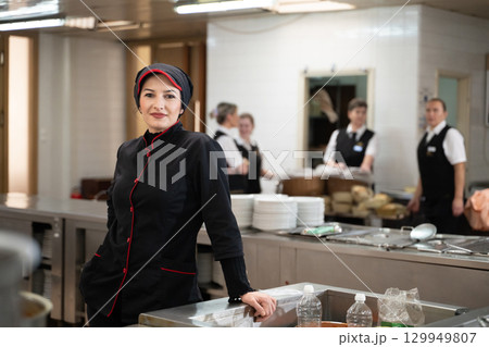 Portrait of a female chef in a black uniform with red details smiling in a professional restaurant kitchen. Image highlights hospitality staff, confidence, and culinary professionalism. 129949807