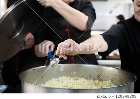 Group of chefs in black uniforms preparing rice in a large pot in a commercial kitchen. Female cooks stirring and adding herbs while working as a team. Concept of teamwork, gastronomy, and hospitality 129949809