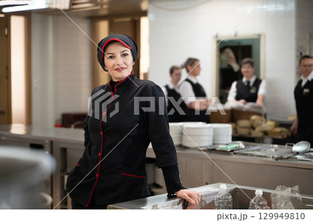 Portrait of a female chef in a black uniform with red details smiling in a professional restaurant kitchen. Image highlights hospitality staff, confidence, and culinary professionalism. 129949810